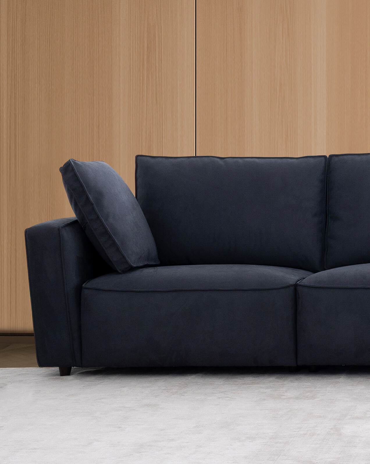 Side view of a dark navy sofa section with two seat cushions and a single back cushion, positioned on a light gray rug in front of a wooden paneled wall.