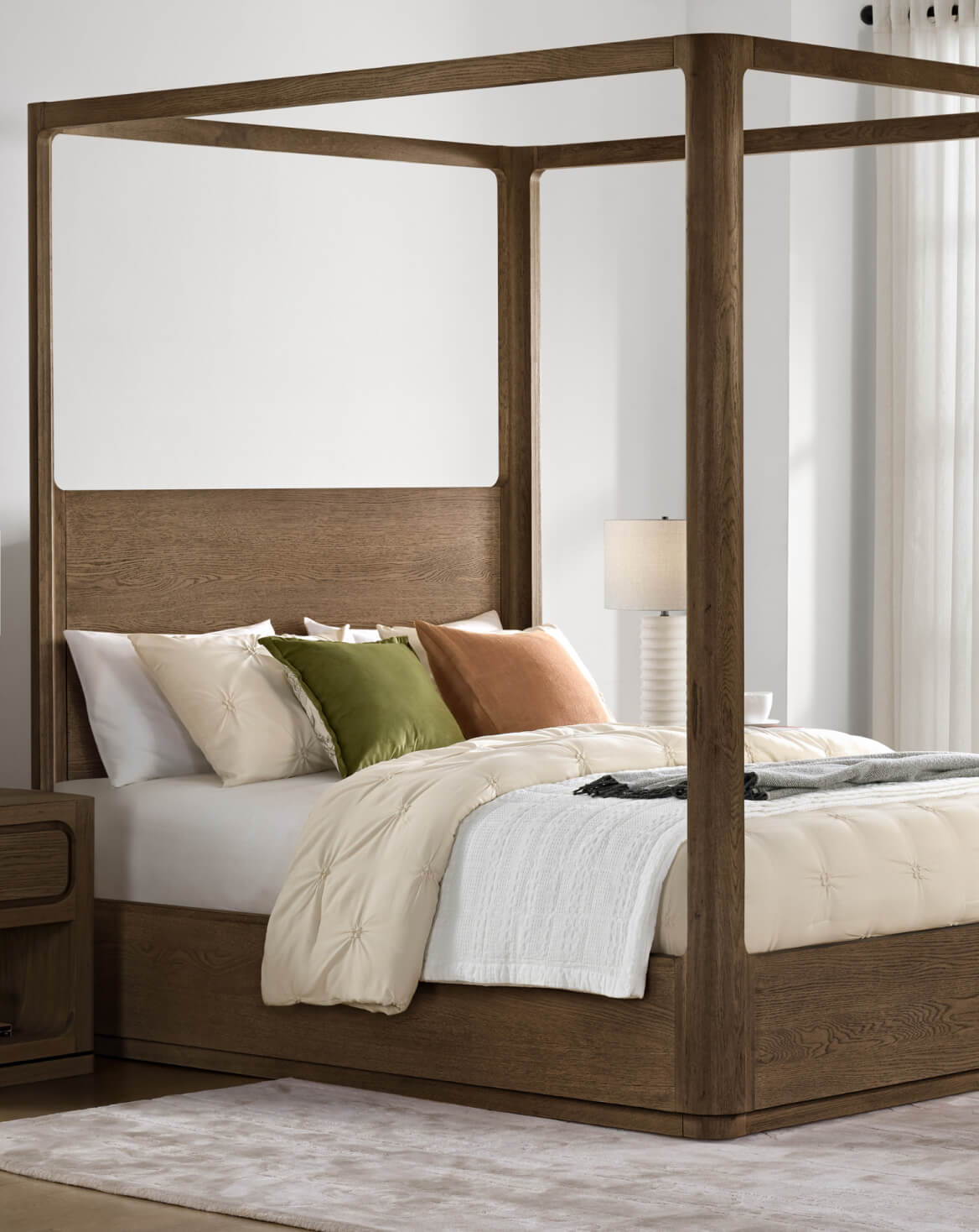 Modern walnut four-poster bed with a solid wood frame and headboard, styled with neutral bedding, accent pillows, a white bedside lamp, and a light rug in a bright, minimalist bedroom.