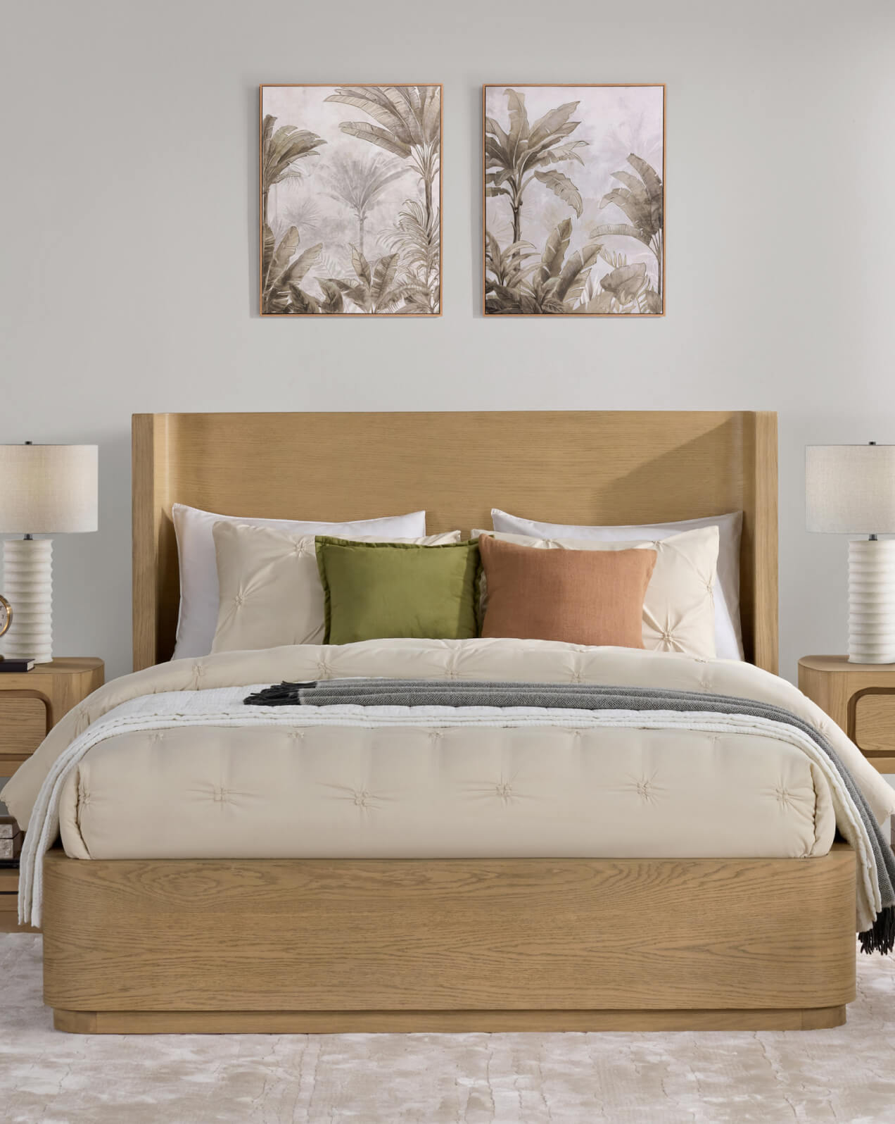 Natural wood panel bed with a wide headboard, dressed in cream bedding with green and terracotta accent pillows, flanked by matching nightstands and lamps, with botanical wall art above.