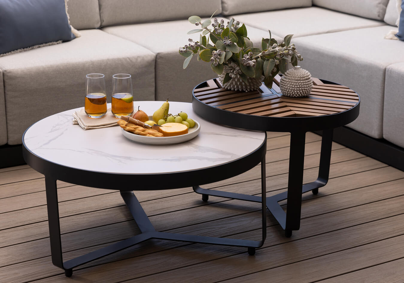 Outdoor coffee tables with marble and wood tops styled with drinks, fruit, and décor beside a grey sofa.